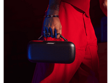 Charger l&#39;image dans la galerie, Personne tenant un haut-parleur Bose noir. Elle porte un pantalon rouge et le tient d&#39;une main.
