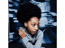 Charger l&#39;image dans la galerie, Une jeune femme porte des écouteurs. Elle a des cheveux bouclés foncés et regarde vers la droite. Un fond bleu flou.
