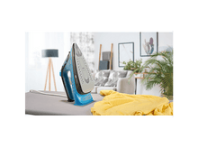 Charger l&#39;image dans la galerie, Un fer à repasser sur une planche à repasser avec un tissu jaune. Un salon est en arrière-plan avec de l&#39;art et des plantes.
