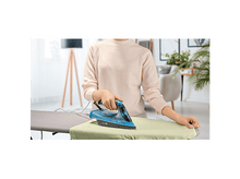 Charger l&#39;image dans la galerie, Une personne repasse un vêtement sur une planche à repasser. Elle tient un fer à repasser bleu sur le vêtement avec un pull rose clair.
