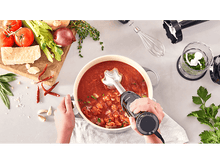 Charger l&#39;image dans la galerie, Une personne mixant de la soupe dans une marmite avec des ingrédients comme des tomates, oignons et herbes.
