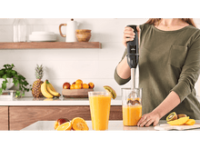 Charger l&#39;image dans la galerie, Une femme dans une cuisine mélangeant une boisson, avec des fruits et un verre de jus sur le comptoir. Étagères en bois.
