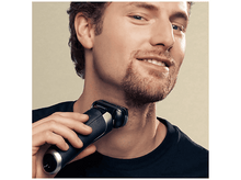 Charger l&#39;image dans la galerie, Un homme se rase le cou avec un rasoir électrique. Il porte un t-shirt noir et sourit.
