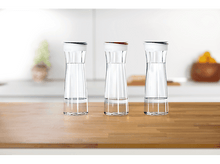 Charger l&#39;image dans la galerie, Trois carafes d&#39;eau transparentes avec des couvercles blancs et des anneaux colorés, sur un comptoir en bois.
