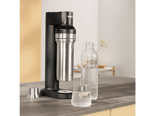 Charger l&#39;image dans la galerie, Un soda maker noir avec une bouteille en acier inoxydable et un verre avec du citron.
