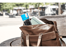 Charger l&#39;image dans la galerie, Un sac en cuir marron avec une bouteille d&#39;eau, un carnet à l&#39;intérieur sur un banc en bois.

