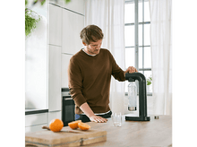 Charger l&#39;image dans la galerie, Un homme en pull marron utilise un fabricant de soda sur un comptoir de cuisine, avec des oranges à proximité.
