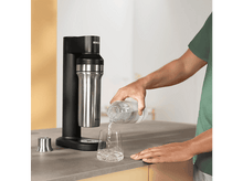 Charger l&#39;image dans la galerie, Personne versant de l&#39;eau d&#39;une bouteille transparente dans un verre, près d&#39;un soda maker, sur un comptoir.
