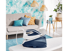 Charger l&#39;image dans la galerie, Un fer à repasser sur une table à repasser, avec du papier peint coloré et un canapé gris en arrière-plan.
