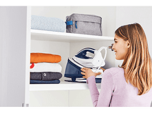 Charger l&#39;image dans la galerie, Une femme range un fer à repasser bleu dans une armoire blanche. Il y a des vêtements pliés et un sac gris sur l&#39;étagère.
