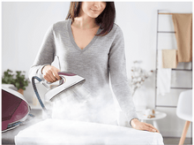 Charger l&#39;image dans la galerie, Une femme repasse une chemise blanche avec un fer à vapeur. La pièce a un mur blanc et une plante.
