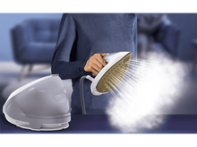 Charger l&#39;image dans la galerie, Une femme tient un fer à repasser à vapeur, d&#39;où s&#39;échappe de la vapeur. Un générateur de vapeur est à gauche.
