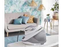 Charger l&#39;image dans la galerie, Un fer à repasser sur une planche à repasser, avec un réservoir d&#39;eau, devant un canapé et déco.
