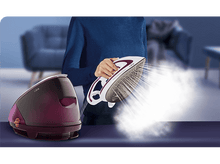 Charger l&#39;image dans la galerie, Femme tenant un fer à repasser à vapeur blanc et violet avec de la vapeur.
