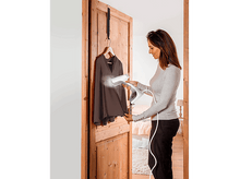 Charger l&#39;image dans la galerie, Une femme qui repasse une chemise marron suspendue à une porte. Elle tient un défroisseur.
