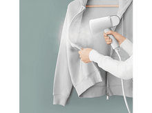 Charger l&#39;image dans la galerie, Une personne défroisse un sweat à capuche gris avec un défroisseur à main blanc. Le mur est bleu-vert.
