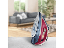 Charger l&#39;image dans la galerie, Fer à repasser rouge et bleu sur une table, arrière-plan flou du salon.
