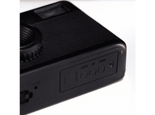 Charger l&#39;image dans la galerie, Gros plan d&#39;un appareil photo noir. Montre une partie de l&#39;objectif, un corps texturé et des boutons de commande. Fond blanc.
