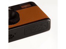 Charger l&#39;image dans la galerie, Un appareil photo noir et marron sur une surface blanche. Il a un objectif, un dessus marron simili cuir et des boutons sur le côté.
