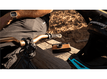 Charger l&#39;image dans la galerie, Une personne avec une montre en or est assise à côté d&#39;un vélo. Un appareil photo est posé sur une surface en bois à côté d&#39;un casque.
