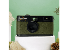 Charger l&#39;image dans la galerie, Un appareil photo Camp Snap vert et noir est posé sur un socle blanc. Le fond est vert avec des accents de pin et de mousse.
