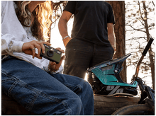 Charger l&#39;image dans la galerie, Une personne tient un appareil photo vert, une autre se tient à proximité. Un casque repose sur une bûche, à côté d&#39;un vélo et d&#39;arbres en arrière-plan.
