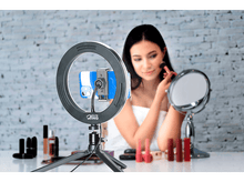 Charger l&#39;image dans la galerie, Une femme se maquille. Une lumière annulaire pour selfies et un miroir sont devant. Rouge à lèvres et autres articles sur la table.
