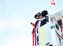 Charger l'image dans la galerie, Pompier dans un ascenseur, regardant sur le côté, tenant un téléphone, ciel bleu en arrière-plan.

