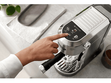 Charger l&#39;image dans la galerie, La main d&#39;une personne appuie sur un bouton d&#39;une machine à expresso argentée. Tasse à café et grains à proximité.
