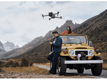 Charger l&#39;image dans la galerie, Un drone plane au-dessus d&#39;une Jeep jaune avec deux personnes, des montagnes en arrière-plan et une route.
