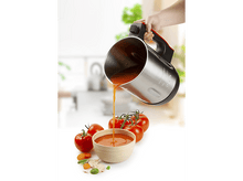 Charger l&#39;image dans la galerie, Une personne verse de la soupe de tomates d&#39;un récipient métallique dans un bol avec des tomates autour.
