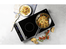 Charger l&#39;image dans la galerie, Une plaque à induction noire avec une poêle de pâtes et une assiette de pâtes, tomates et pain.
