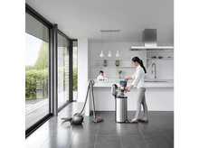 Charger l&#39;image dans la galerie, Une femme dans une cuisine blanche jette des ordures dans une poubelle argentée.
