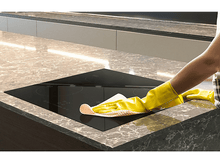 Charger l&#39;image dans la galerie, Une personne portant des gants jaunes nettoie une table de cuisson noire dans une cuisine.

