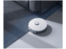 Charger l&#39;image dans la galerie, Aspirateur robot blanc sur un tapis bleu. Un canapé gris est visible en arrière-plan.
