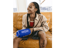 Charger l&#39;image dans la galerie, Femme aux dreadlocks souriante tenant un haut-parleur bleu. Elle est sur un canapé orange, portant une veste et un short.
