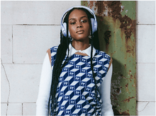 Charger l&#39;image dans la galerie, Une femme aux tresses noires porte un casque et un gilet à motifs devant un mur.
