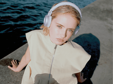 Charger l&#39;image dans la galerie, Une femme avec un casque violet clair est assise sur une corniche près de l&#39;eau. Elle porte une tenue beige et regarde la caméra.
