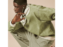 Charger l&#39;image dans la galerie, Homme en veste et pantalon verts. Écouteurs gris et écharpe blanche.
