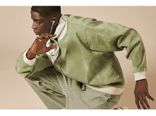 Charger l&#39;image dans la galerie, Homme en veste verte et pantalon rayé, portant des écouteurs. Il est accroupi.
