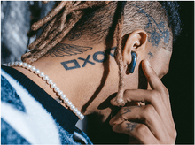 Charger l&#39;image dans la galerie, Homme avec des tatouages sur le cou et un écouteur dans l&#39;oreille. Collier de perles. Fond sombre. Pull noir et blanc.
