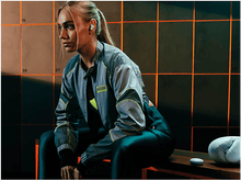 Charger l'image dans la galerie, Femme blonde portant des écouteurs dans une salle de sport. Gants de boxe et casiers en arrière-plan.
