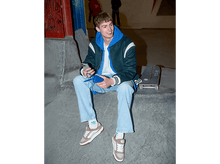 Charger l'image dans la galerie, Jeune homme en sweat à capuche bleu assis sur une rampe avec un skateboard. Il tient un téléphone et un chargeur.
