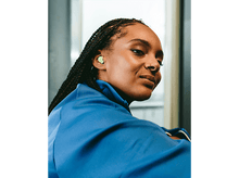 Charger l'image dans la galerie, Femme aux cheveux noirs tressés, portant une veste bleue et des écouteurs jaune-vert, regardant la caméra.
