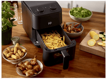 Charger l&#39;image dans la galerie, Friteuse à air noire avec des frites. Des bols de nourriture entourent la friteuse. Une plante et une surface en bois.
