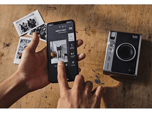 Charger l&#39;image dans la galerie, Mains d&#39;une personne tenant un téléphone affichant une photo, avec un appareil photo et des photos sur une table.
