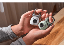 Charger l&#39;image dans la galerie, Deux petites caméras dans les mains. Une blanche, une verte. Les deux ont une boucle.
