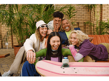 Charger l&#39;image dans la galerie, Groupe de cinq amis divers riant ensemble, assis sur un canapé avec un skateboard.
