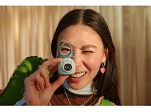 Charger l&#39;image dans la galerie, Une femme fait un clin d&#39;œil en tenant un petit appareil photo vert et bleu. Elle porte des boucles d&#39;oreilles et un collier de perles.
