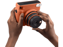 Charger l&#39;image dans la galerie, Une personne tient un appareil photo Instax orange sur un fond blanc.
